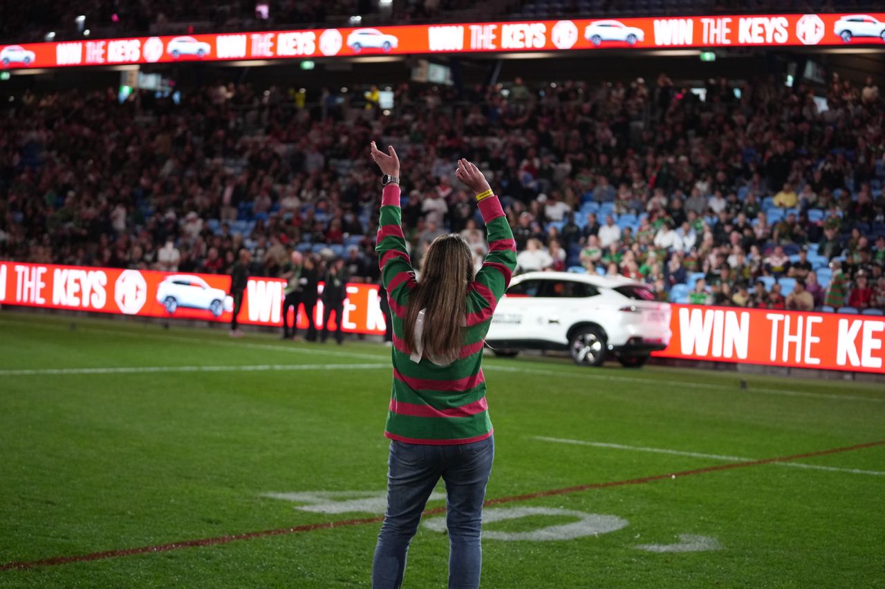 A fan in a Rabbitohs jersey with arms raised on the field at Allianz Stadium, the MG car and the Win the Keys boards behind
